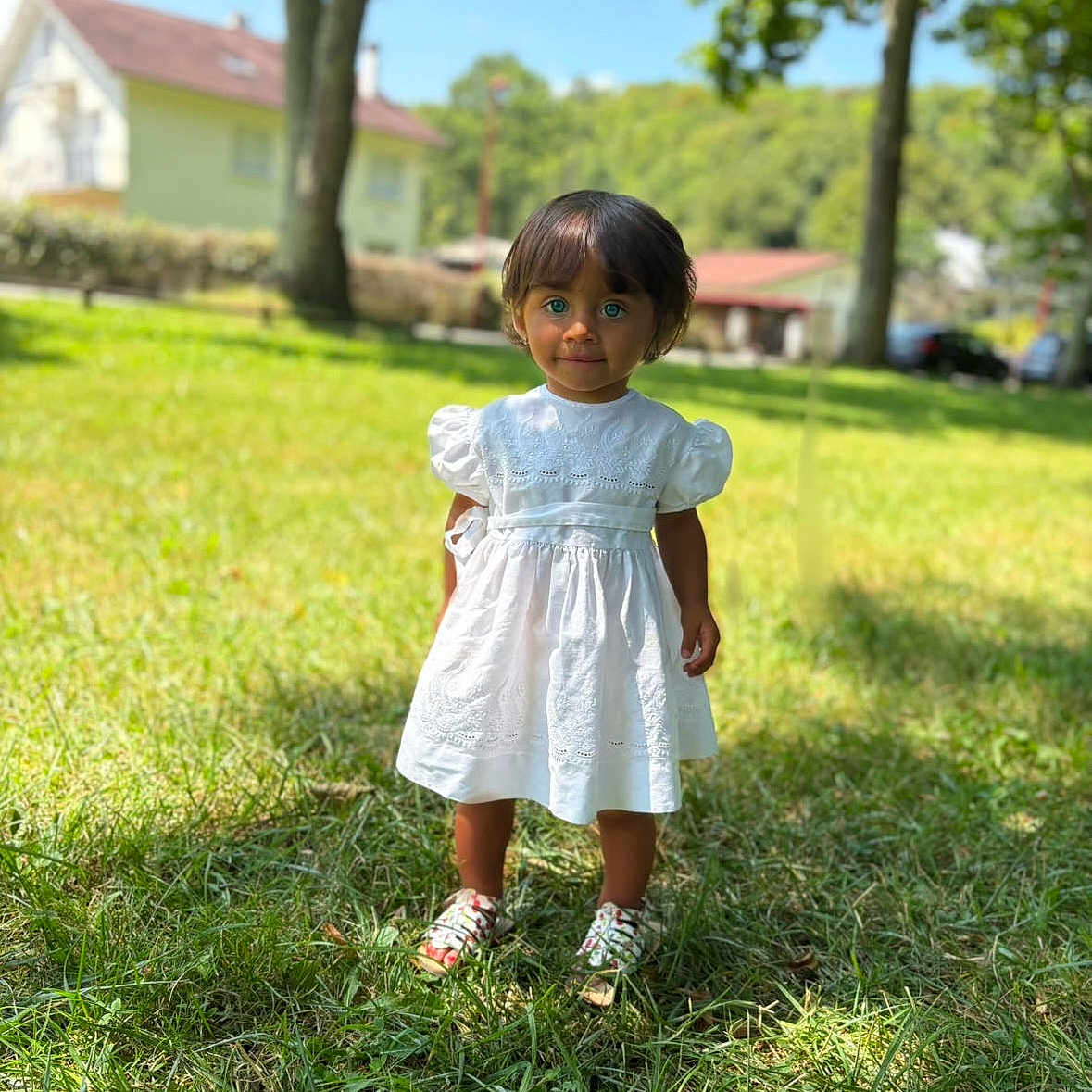Calyana participe au concours pour gagner de l'argent avec cette photo : background_blur, blue_eyes, child, cute, daylight, grass, greenery, house, nature, outdoor, person, portrait, shoes, smiling, standing, sunny, toddler, trees, white_dress, young_child