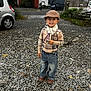 boots, car, casual, child, cloudy_sky, daytime, fashion, flat_cap, gravel, happy, industrial_building, jeans, outdoor, person, plaid_sweater, scarf, smiling, standing, street, urban