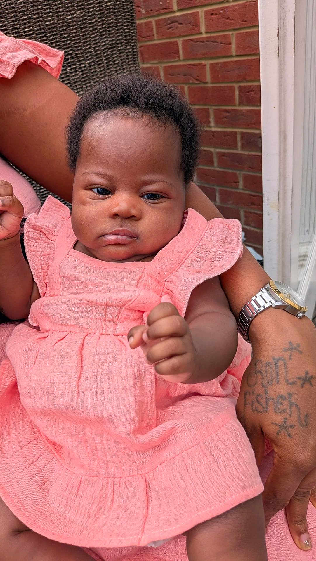 Princess is registered to the contest to win money with this photo: baby, infant, pink_dress, clothing, person, arm, hand, tattoo, watch, bracelet, skin, face, expressionless, closeup, outdoor, brick_wall, fabric, sitting, holding, adult
