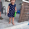 child, toddler, girl, polka_dot_dress, barefoot, smiling, toy, porch, wooden_floor, outdoor, happy, person, short_hair, blue_dress, playful, daylight, home, fence, door, window