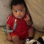 Navy is registered to the contest to win money with this photo: baby, child, red_clothing, sneakers, pacifier_clip, couch, indoor, cute, infant, portrait, relaxed, curly_hair, looking_at_camera, seated, casual, adorable, young_child, expression, comfortable, furniture