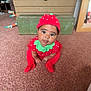 baby, child, strawberry_costume, red_clothing, green_collar, hat, carpet, indoor, furniture, wooden_chest, lamp, toy, curious_expression, crawling, cute, person, floor, face, headwear, toddler