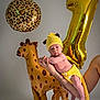 baby, infant, balloon, giraffe_balloon, number_one_balloon, gold_balloon, leopard_balloon, party, celebration, yellow_hat, knit_hat, crochet_outfit, diaper_cover, adult_hand, arm, studio_photography, portrait, gray_background, neutral_expression, cute
