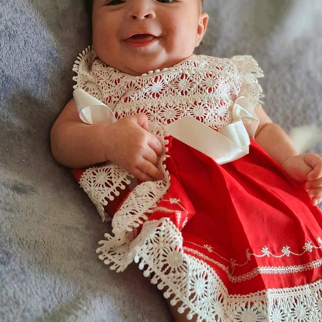 Liara is registered to the contest to win money with this photo: baby, smiling, red_dress, headband, flower, lace, blanket, lying_down, infant, cute, happy, child, portrait, soft_texture, indoors, young, person, skin, hand, foot