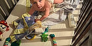 Malakai is registered to the contest to win money with this photo: baby, crib, blanket, diaper, toy, bottle, floor, wood, light, shadow, striped_towel, plush_toy, colorful_toy, person, infant, looking_up, indoor, soft_toy, bed, sleeping_area