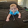 baby, blue_clothing, bed, black_bedspread, charging_cable, child, curious, daylight, face, furniture, indoor, lying_down, person, remote_control, room, window, window_blinds, young_child, hands, head