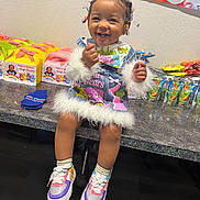 Elaya is registered to the contest to win money with this photo: child, toddler, smiling, birthday, party, presents, gift_boxes, colorful, portrait, sitting, indoors, playroom, braids, hair_beads, dress, fur_trim, sneakers, juice_pouches, cartoon_decor, happy
