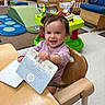 Mia joined the competition — help win amazing prizes! toddler, child, high_chair, book, smile, indoor, nursery, daycare, play_area, toy, wooden_furniture, flooring, happy, person, clothing, hair, table, seat, reading, playtime