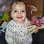 toddler, child, smiling, toy, giraffe, curly_hair, indoor, play_area, stuffed_animals, colorful, happy, person, clothing, long_sleeve_shirt, patterned_shirt, pacifier_clip, black_pants, floor, home, cute