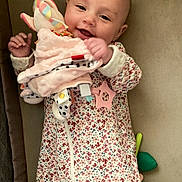 Rowan is registered to the contest to win money with this photo: baby, infant, smile, toy, onesie, floral_pattern, plush, hand, face, portrait, lying_down, couch, indoor, happy, teether, close_up, skin, head, chubby_cheeks, nursery