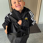 Mylan participe au concours pour gagner de l'argent avec cette photo : child, boy, smiling, indoor, door, floor, bag, black_clothing, tracksuit, shoes, happy, standing, portrait, young, sporty, casual, person, accessory, holding, expression