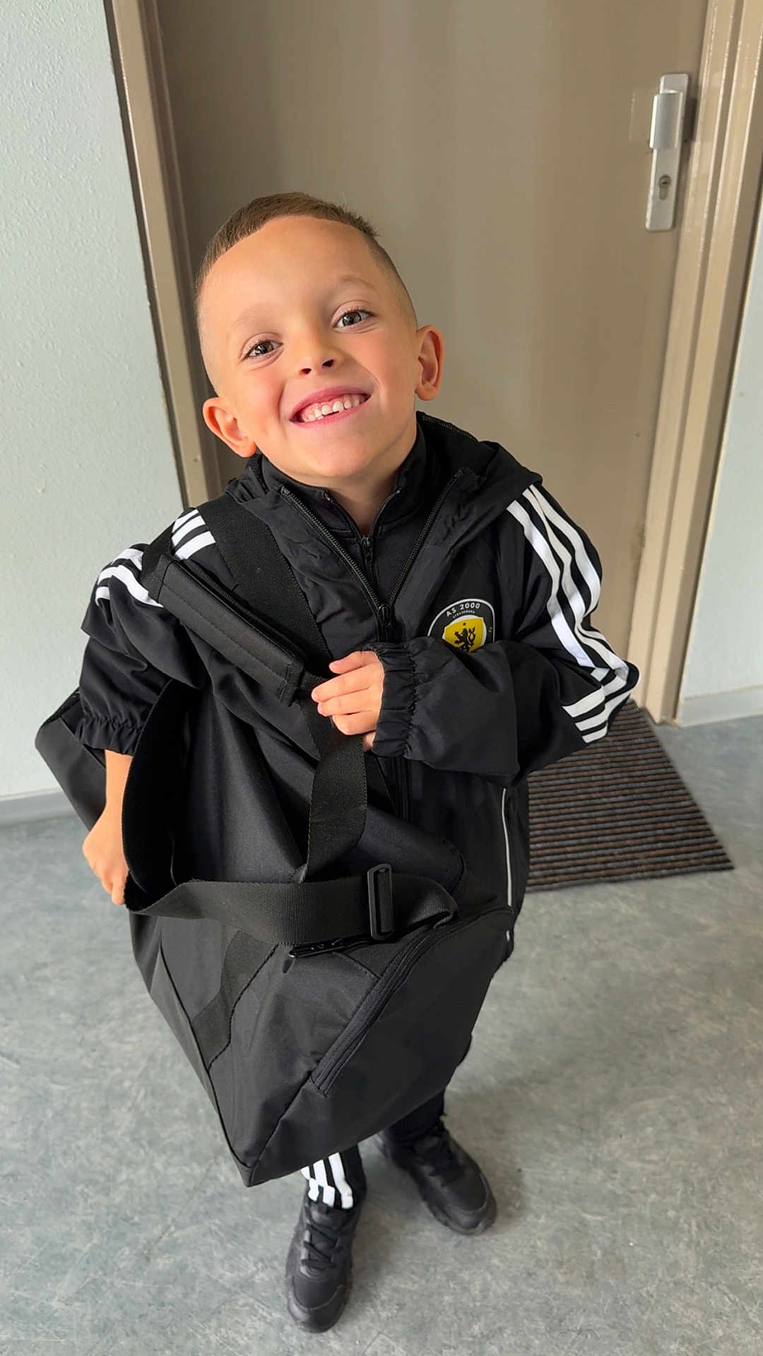 Mylan participe au concours pour gagner de l'argent avec cette photo : child, boy, smiling, indoor, door, floor, bag, black_clothing, tracksuit, shoes, happy, standing, portrait, young, sporty, casual, person, accessory, holding, expression