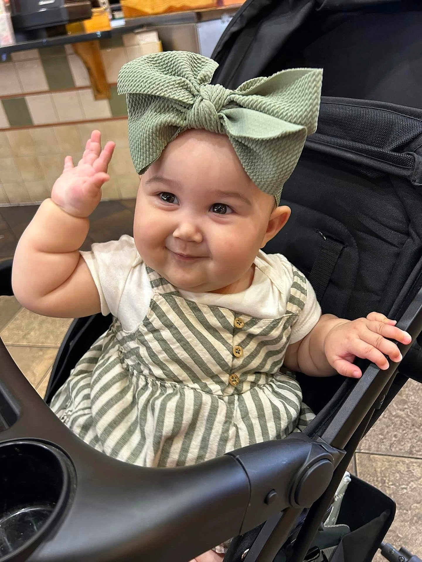 Sutton is registered to the contest to win money with this photo: baby, child, smiling, waving, bow, headwear, stroller, seat, striped_dress, clothing, cute, happy, indoor, floor_tiles, person, infant, cheeks, hand, portrait, playful