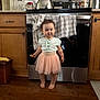toddler, child, kitchen, barefoot, tutu, pink_skirt, mint_top, wood_floor, oven, kitchen_towel, teapot, cup, cabinet, snack, smiling, standing, indoor, appliance, curly_hair, playful