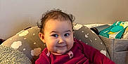 Sutton is registered to the contest to win money with this photo: baby, child, smiling, curly_hair, long_sleeve_shirt, maroon_clothing, cushion, blanket, diaper, indoor, cozy, infant, person, cute, plush, seated, happy, soft_texture, home, relaxed