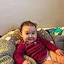 baby, child, smiling, curly_hair, long_sleeve_shirt, maroon_clothing, cushion, blanket, diaper, indoor, cozy, infant, person, cute, plush, seated, happy, soft_texture, home, relaxed