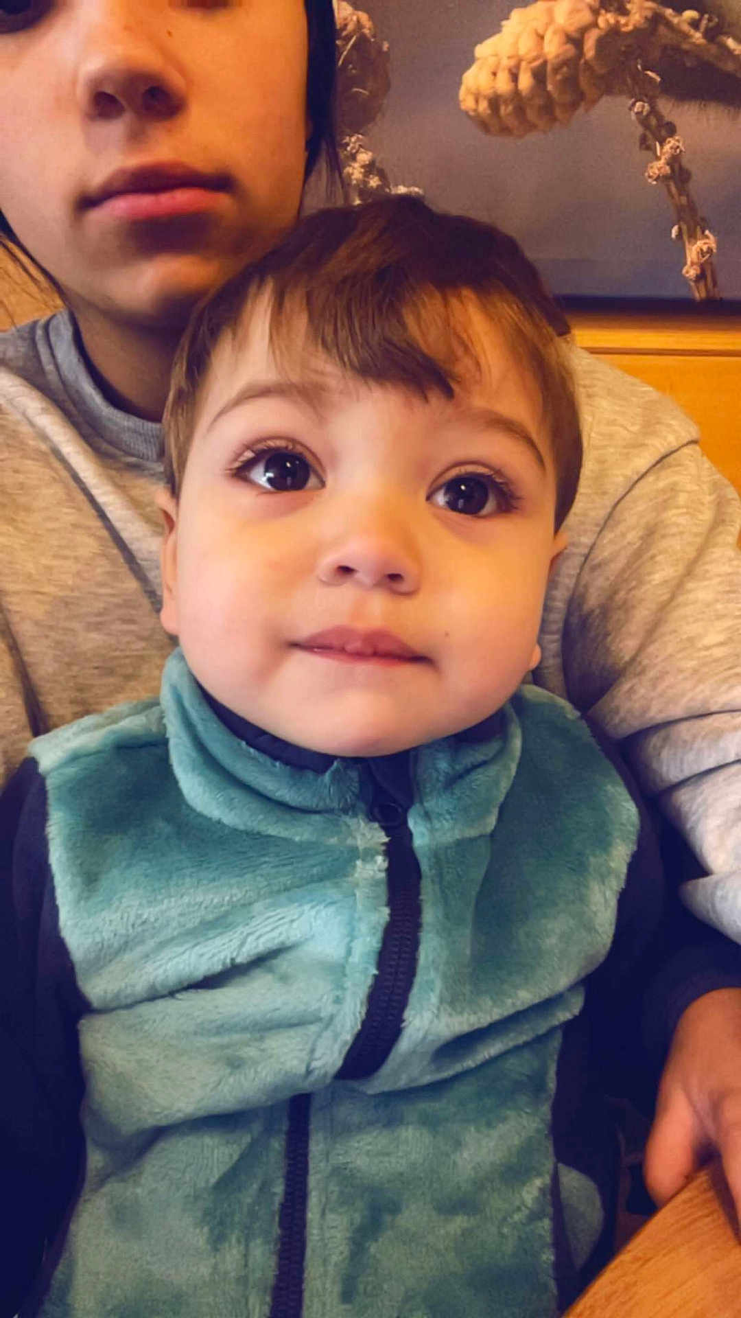 Aaron participe au concours pour gagner de l'argent avec cette photo : child, toddler, person, adult, selfie, portrait, close_up, big_eyes, fleece_jacket, teal_jacket, face, eyelashes, hair, indoor, wooden_table, hand, sitting, expression_curious, background_art, casual_clothing