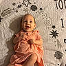 baby, infant, child, smiling, happy, portrait, lying_down, blanket, bed, dress, orange_clothing, crossed_legs, barefoot, face, eyes, open_mouth, indoor, warm_lighting, star_pattern, photography