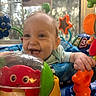 Esme is registered to the contest to win money with this photo: baby, child, infant, smiling, laughing, happy, portrait, indoor, nursery, activity_center, baby_toys, playtime, colorful, orange_toy, red_toy, green_toy, seated, sunlight, window, close_up