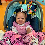 Kalaya is registered to the contest to win money with this photo: baby, car_seat, blanket, pink, toy, smile, child, person, indoor, clothing, face, head, seat, colorful, happy, holding, fabric, pattern, hand, background