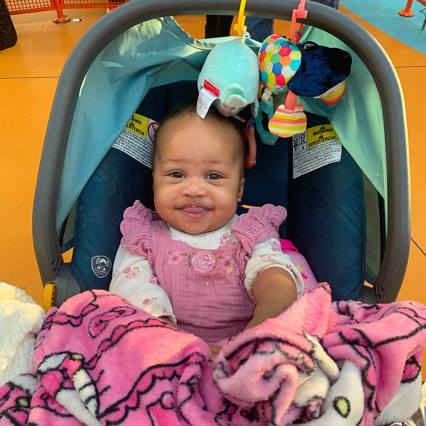 Kalaya is registered to the contest to win money with this photo: baby, car_seat, blanket, pink, toy, smile, child, person, indoor, clothing, face, head, seat, colorful, happy, holding, fabric, pattern, hand, background