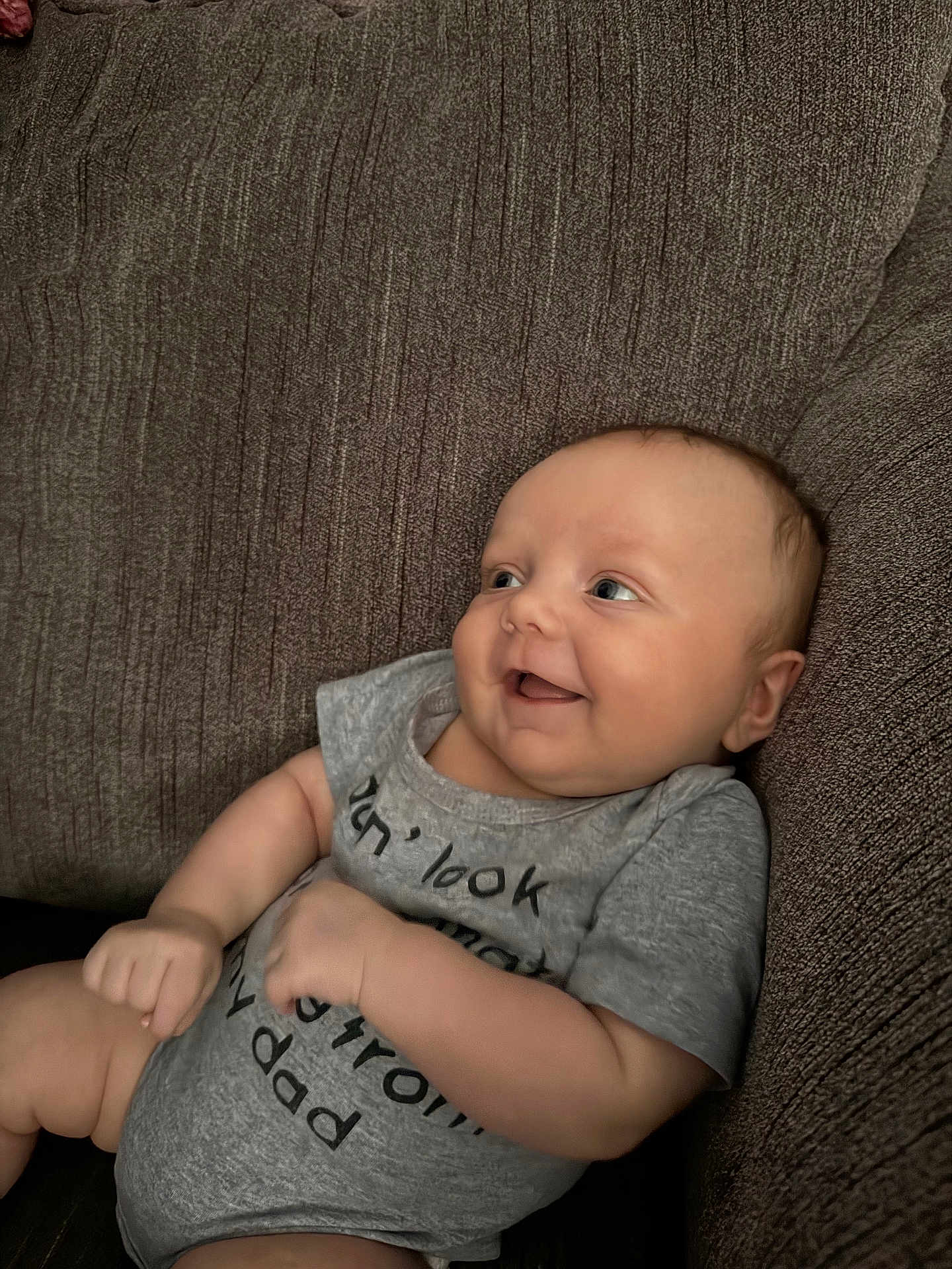 Bellamy is registered to the contest to win money with this photo: baby, infant, smile, couch, gray_onesie, happy, face, arm, hand, skin, textile, home, relaxation, seated, cute, child, indoors, portrait, person, cozy