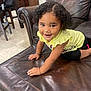 toddler, child, smiling, curly_hair, yellow_shirt, black_shorts, leather_ottoman, crawling, indoor, living_room, furniture, happy, playful, person, floor, skin, hand, head, eyes, cute