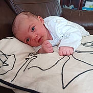 Evelyn is registered to the contest to win money with this photo: baby, infant, cushion, pattern, brown_leather, couch, indoor, person, clothing, white_outfit, face, hand, fist, head, newborn, resting, looking, young_child, skin, expression