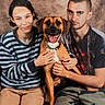 dog, person, smile, indoor, portrait, casual_clothing, striped_sweater, camo_pants, friendship, pet, brown_dog, happy, seated, hands, face, short_hair, long_hair, wall_background, warm_lighting, looking_at_camera