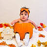 baby, infant, child, pumpkin_costume, orange, autumn, fall_leaves, headband, bow, sitting, portrait, indoor, quilt, blanket, white_socks, seasonal, five_months, decorative_sign, wide_eyes, smiling