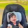 baby, child, infant, stroller, smiling, laughing, outdoor, park, portrait, face, hair_bow, pink_bow, dress, harness, seatbelt, carriage, sunlight, grass, sidewalk, happy