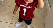 Noah is registered to the contest to win money with this photo: child, toddler, smiling, happy, portrait, football_jersey, red_hat, baseball_cap, sportswear, arizona, nfl, number_one, indoors, hallway, tile_floor, standing, shoes, white_sneakers, casual_clothing, hand_on_hat