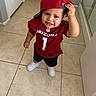 child, toddler, smiling, happy, portrait, football_jersey, red_hat, baseball_cap, sportswear, arizona, nfl, number_one, indoors, hallway, tile_floor, standing, shoes, white_sneakers, casual_clothing, hand_on_hat