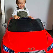 Menzo participe au concours pour gagner de l'argent avec cette photo : toddler, child, toy_car, red, audi, indoor, smiling, steering_wheel, flooring, overalls, white_shirt, head, hair, eyes_closed, playtime, vehicle, license_plate, front_view, mirror, happy