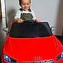 toddler, child, toy_car, red, audi, indoor, smiling, steering_wheel, flooring, overalls, white_shirt, head, hair, eyes_closed, playtime, vehicle, license_plate, front_view, mirror, happy