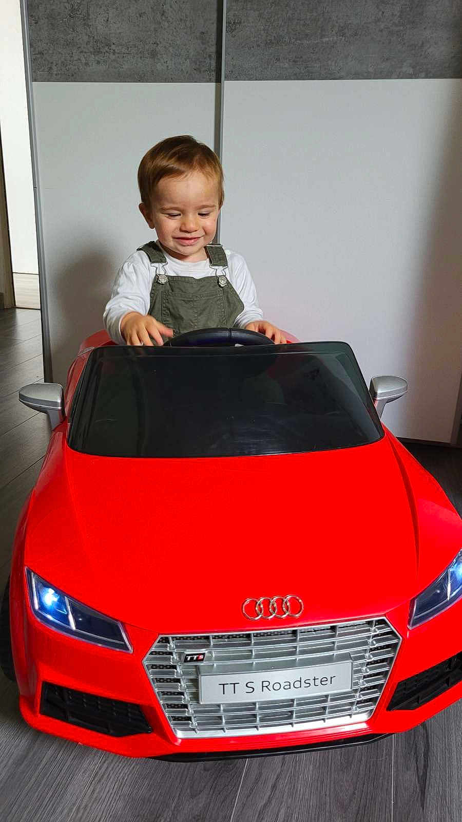 Menzo participe au concours pour gagner de l'argent avec cette photo : toddler, child, toy_car, red, audi, indoor, smiling, steering_wheel, flooring, overalls, white_shirt, head, hair, eyes_closed, playtime, vehicle, license_plate, front_view, mirror, happy