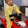 toddler, child, toy, atv, red, indoor, yellow_clothing, floor, furniture, person, vehicle, play, young_child, expression, handlebars, casual, home, footwear, curly_hair, portrait