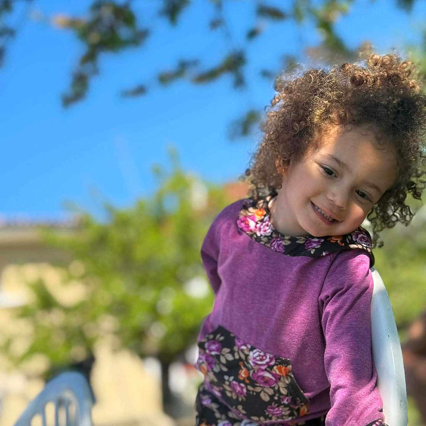 Lunna a rejoint le concours — aidez-le/la à gagner de superbes lots ! child, curly_hair, purple_clothing, floral_pattern, outdoor, blue_sky, greenery, smiling, white_chair, playful, sunlight, portrait, casual_wear, nature, happy, young_child, daylight, person, fashion, background_blur