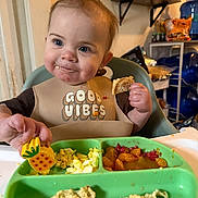 Fiona is registered to the contest to win money with this photo: baby, bib, child, cracker, eating, face, food, fruit, happy, high_chair, indoor, kitchen, meal, messy, person, pineapple_toy, plate, scrambled_eggs, smiling, toy