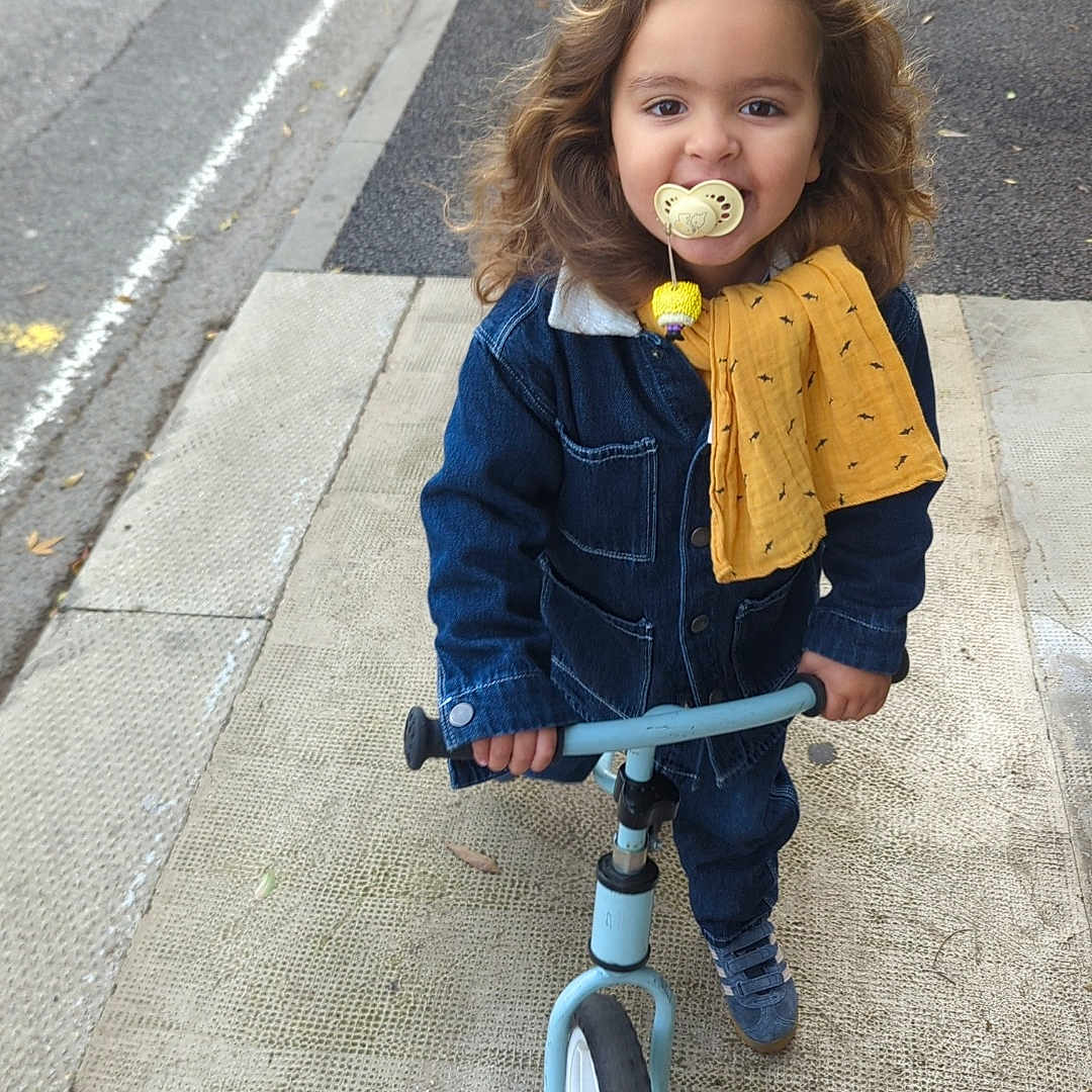 Isaac participe au concours pour gagner de l'argent avec cette photo : balance_bike, casual_clothing, child, curly_hair, daytime, denim_jacket, footwear, happy, outdoor, pacifier, person, play, sidewalk, smiling, street, toddler, toy, urban, yellow_cloth, young_child