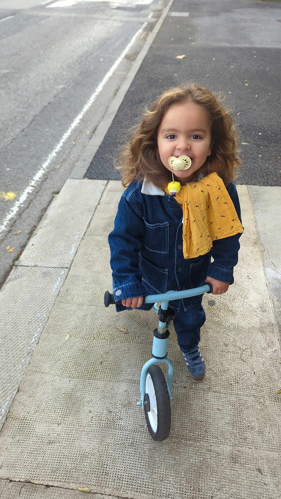 Isaac participe au concours pour gagner de l'argent avec cette photo : child, toddler, balance_bike, sidewalk, denim_jacket, pacifier, yellow_cloth, curly_hair, outdoor, street, footwear, casual_clothing, happy, person, urban, play, toy, young_child, smiling, daytime