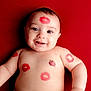 arm, baby, chest, child, closeup, cute, expression, face, happy, infant, kiss_mark, lipstick, lying_down, person, playful, portrait, red_background, skin, smiling, tongue