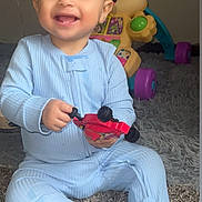 Mikah joined the competition — help win amazing prizes! baby, child, smiling, toy_car, onesie, carpet, indoor, playtime, happy, cute, infant, plastic_toy, floor, sitting, holding, expression, childhood, person, young_child, playful