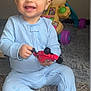 baby, child, smiling, toy_car, onesie, carpet, indoor, playtime, happy, cute, infant, plastic_toy, floor, sitting, holding, expression, childhood, person, young_child, playful