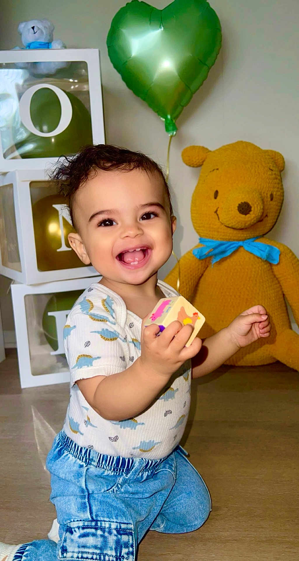 Mikah is registered to the contest to win money with this photo: toddler, child, smiling, indoor, toy, block, knitted_bear, balloon, green_balloon, blue_clothing, denim_pants, happy, cute, playful, floor, home, person, face, baby, joy