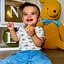 toddler, child, smiling, dinosaur_print, shirt, blue_jeans, socks, toy_block, balloons, green_balloons, birthday, celebration, yellow_teddy_bear, stuffed_animal, ribbon, floor, indoor, happy, face, person