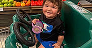Judah is registered to the contest to win money with this photo: toddler, child, shopping_cart, grocery_store, produce, tomatoes, avocados, onions, snack_container, smiling, black_shirt, blue_shorts, green_cart, race_car_design, indoor, supermarket, food, happy, person, retail