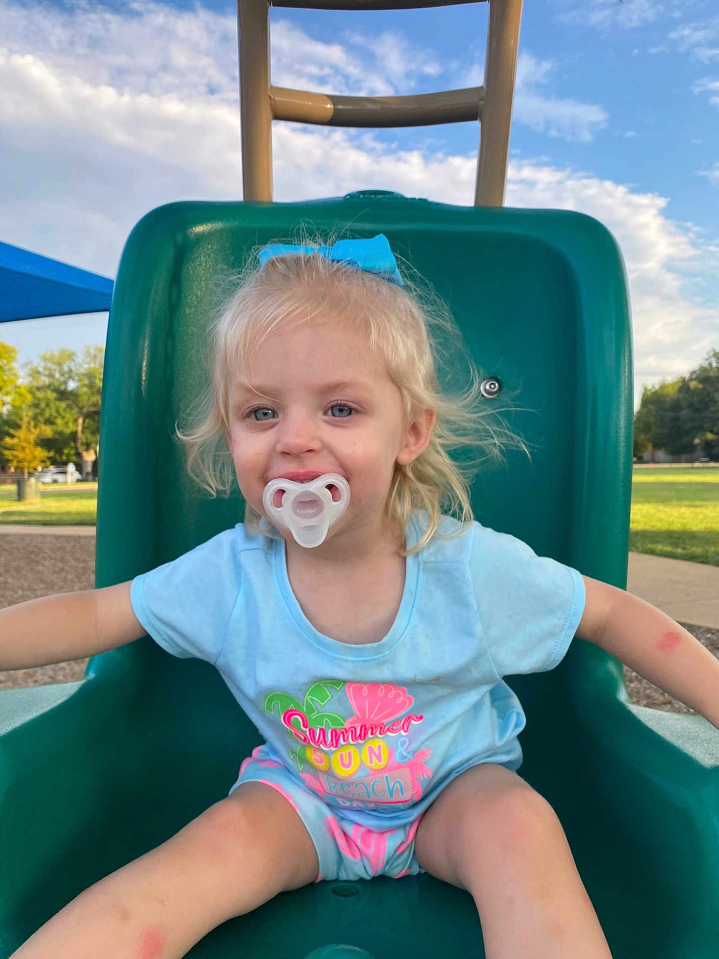 Ellie joined the competition — help win amazing prizes! child, toddler, pacifier, slide, playground, outdoor, blue_sky, summer_clothing, bow, grass, park, happy, smiling, casual, daylight, shorts, tshirt, play, young_child, fun