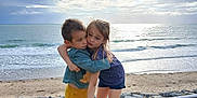 Kaly a rejoint le concours — aidez-le/la à gagner de superbes lots ! child, children, hug, beach, ocean, waves, sky, clouds, bench, sand, rocks, barefoot, casual_clothing, outdoor, friendship, affection, nature, sunlight, toddler, emotion