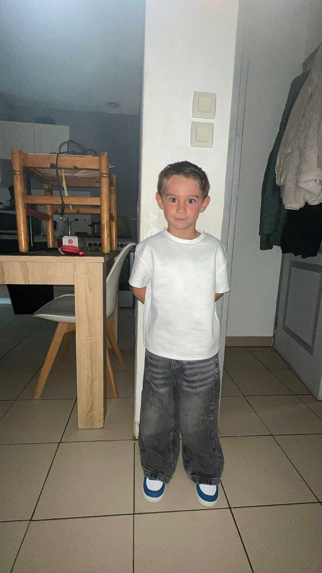 Iago participe au concours pour gagner de l'argent avec cette photo : child, boy, indoor, white_tshirt, jeans, shoes, tile_floor, chair, table, wooden_furniture, door, coat, light_switch, expression, standing, mischievous, home, casual_clothing, person, portrait
